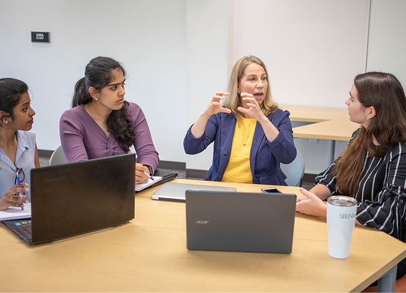 Three female students listen to a professor in a small group setting.