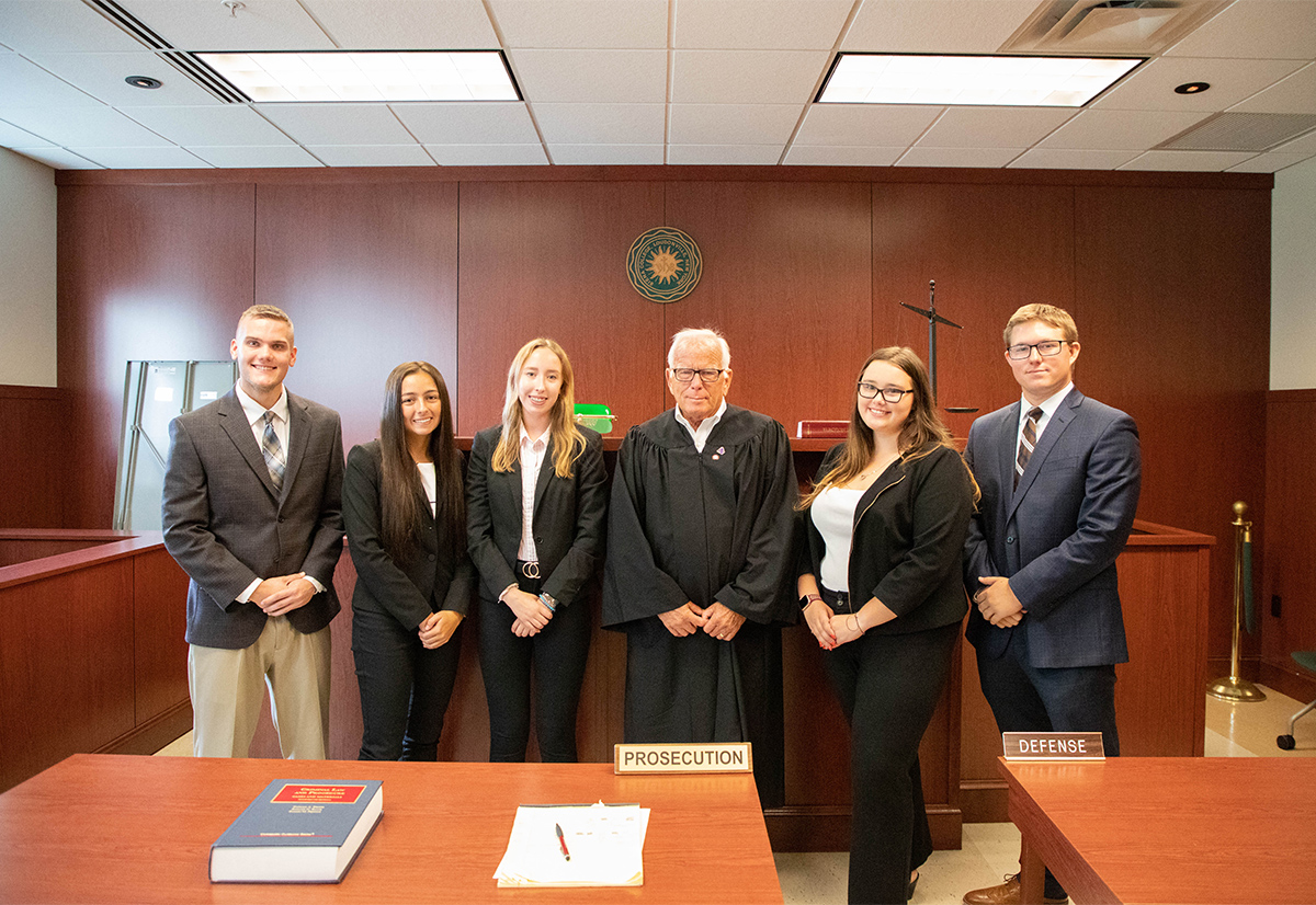 A group of students pose with a judge in robes in a courtroom.
