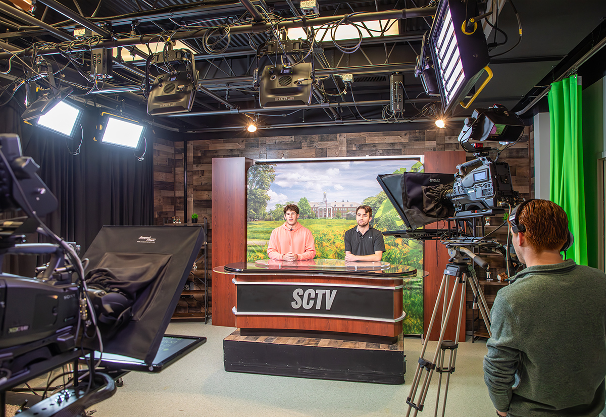 Two male students sit a news desk in a television studio on campus.