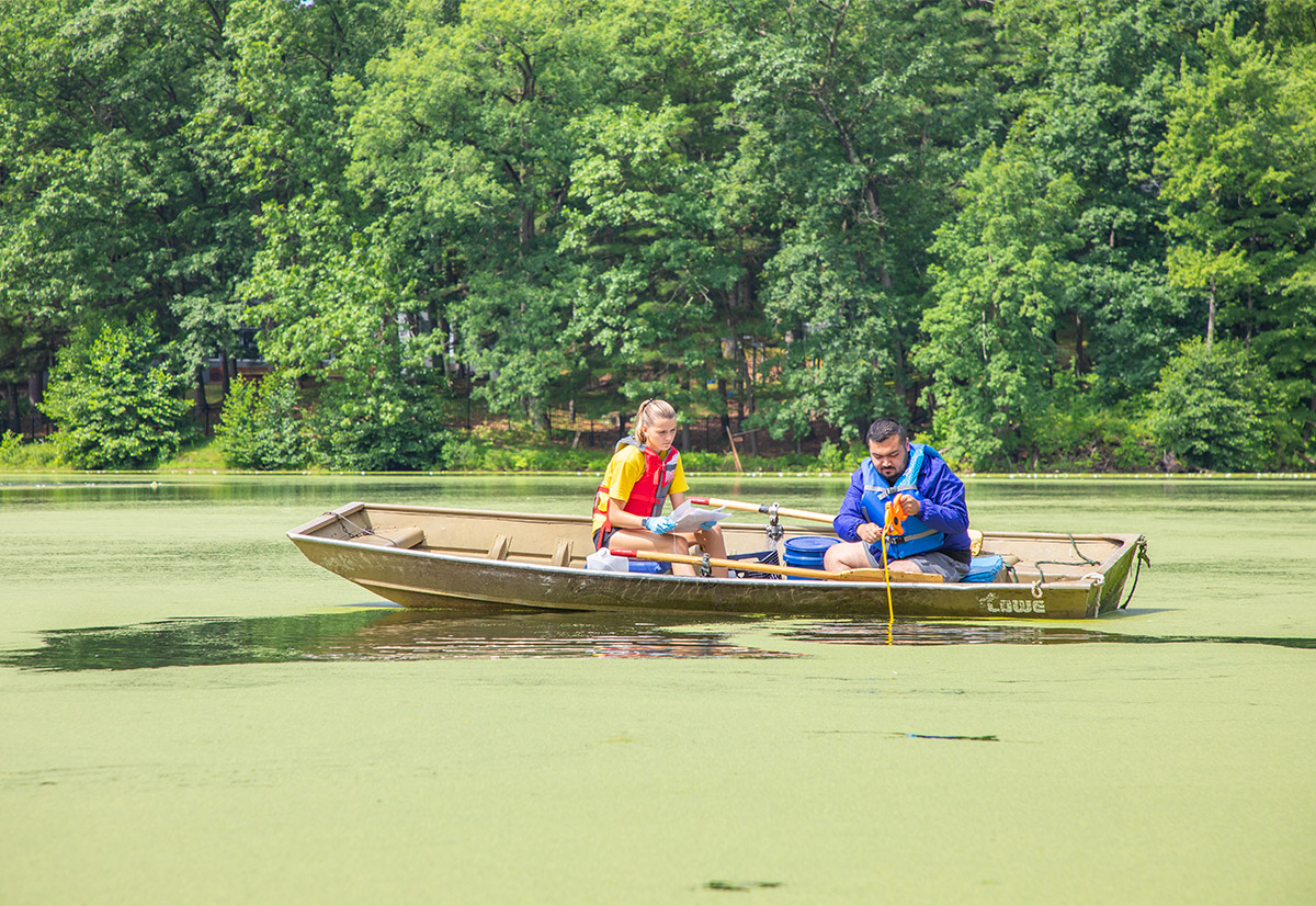 Two students collect water samples in a canoe on a green pond.