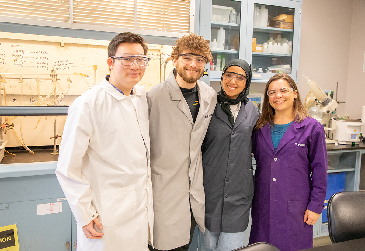 Three students in grey lab coats pose with a professor in a purple lab coat in a science lab on campus.